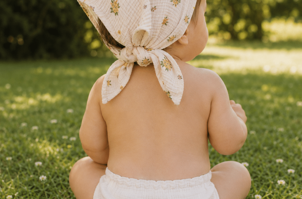 Bébé assis dans l’herbe avec un lange fleuri noué sur la tête pour se protéger du soleil