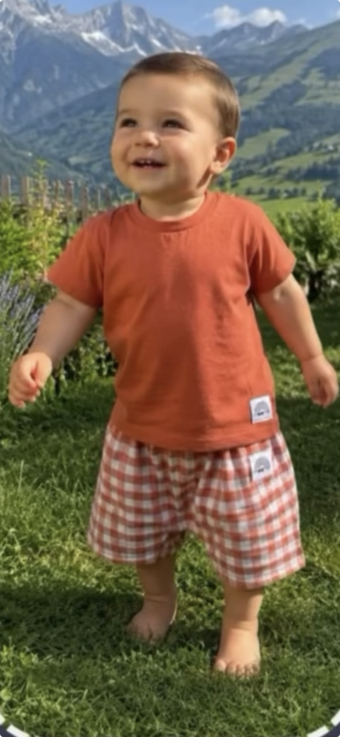 Bébé souriant portant un short vichy terracotta et blanc et un t-shirt terracotta dans l’herbe en été